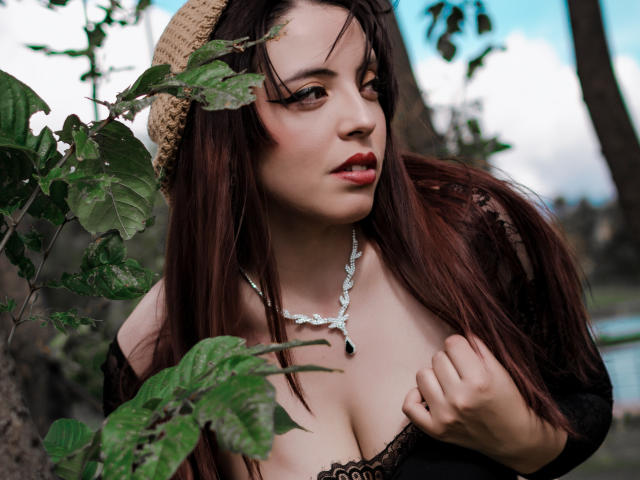 Rosalinnes with brown hair, wearing a straw hat, black lace top, showing her large tits, silver necklace, and red lipstick, standing near green leaves, looking to the side.
