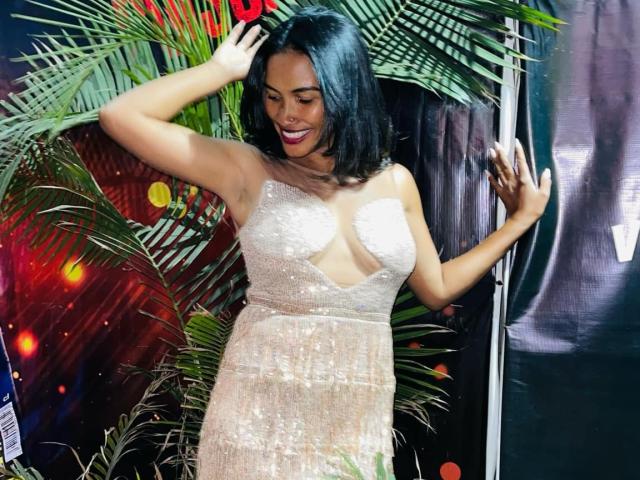Finoana with dark hair, medium tits, wearing a sparkly, one-shoulder dress, hands up, smiling, standing in front of green palm leaves. Her ass is slightly visible.