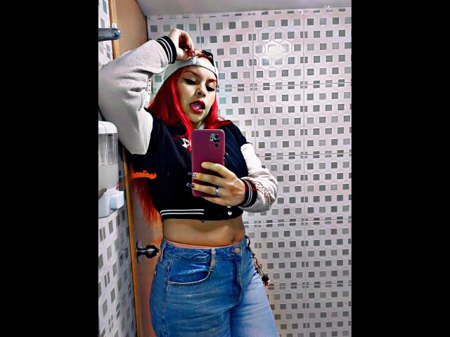 AlanaRoth with red hair, wearing a black and white varsity jacket, exposing her midriff, and blue jeans, taking a selfie in a bathroom, showing off her tits.