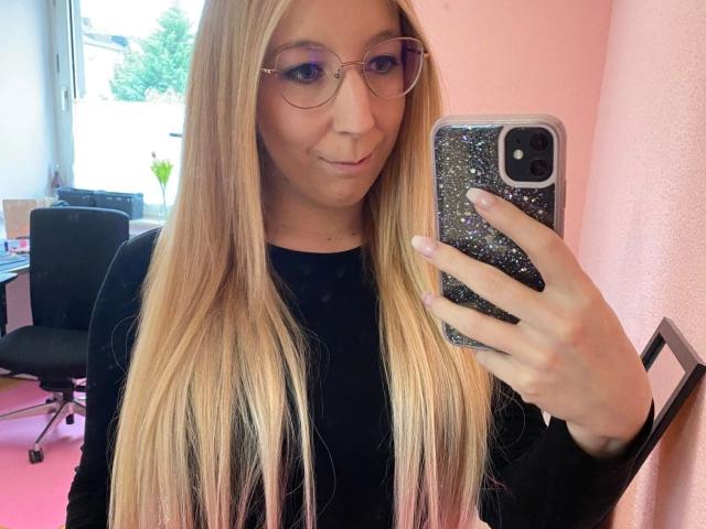 JustKerry-hot with long blonde hair, glasses, and a black top, taking a selfie with a glittery phone case, showing a slight smile. Boobs are not visible.