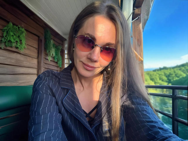 LucieMack with long brown hair, wearing pink-tinted sunglasses and a pinstriped blue shirt, sitting on a green bench, smiling slightly, with a wooded landscape in the background.