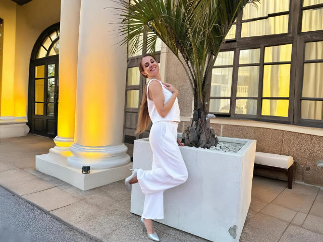 LucieMack leans against a white planter, wearing a white sleeveless top and high-waisted pants, with her hand on her chest, smiling, and her ass slightly raised.