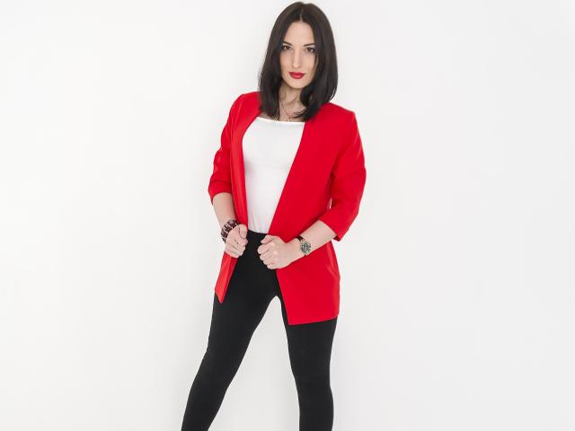MillaXQueen with dark hair, red open cardigan, white top, black pants, red lipstick, tattoo on right wrist, standing, hands near waist.