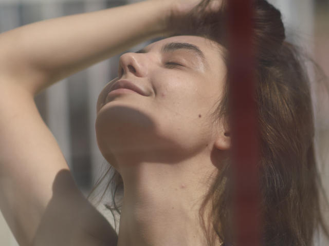 Le visage de CassieRose, les yeux fermés, les bras levés, cheveux bruns, léger sourire, petite tache de naissance sur la joue gauche, peau illuminée par le soleil, ombre douce, ligne verticale rouge au premier plan..