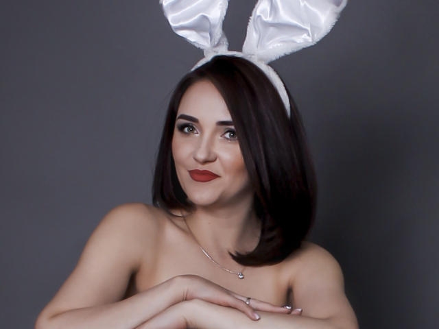 VeronicaLaLux with dark brown bob, white bunny ears, red lipstick, and silver necklace, arms crossed, showing bare shoulders and small, perky tits. Gray background.