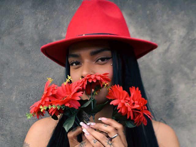 EmmaEbony with long black hair, wearing a red hat, holding red chrysanthemums to her face, showing tattoos on her fingers, and wearing a nose ring.