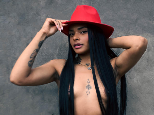 EmmaEbony, com cabelo preto longo, chapéu vermelho e tatuagens, sem blusa, mostrando seios pequenos, braços levantados, fundo de parede cinza texturizado, batom rosa e um colar com um coração..