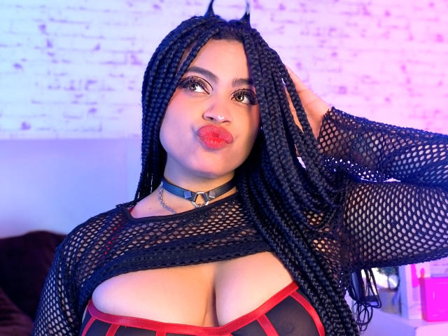 AprilKershaw with long black braids, wearing a black fishnet top, red and black bustier, and black choker, pouting with red lipstick, hand in hair. Visible tits, cute.