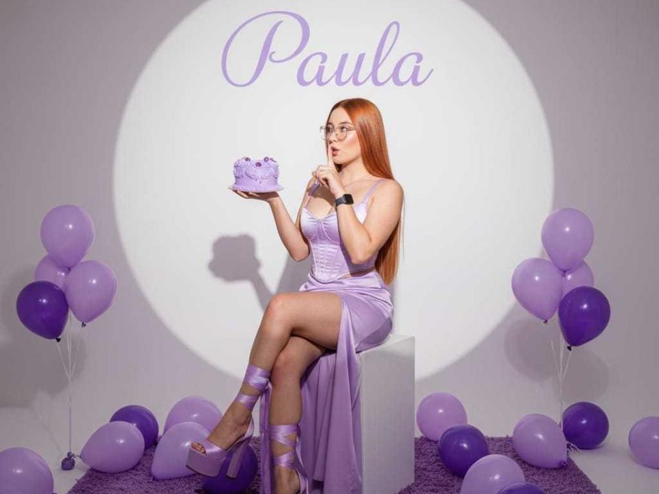 MaddyMe met lang rood haar, een bril, een paarse jurk, hoge hakken en een lint, die een paarse taart vasthoudt, omringd door paarse ballonnen, zittend op een witte kubus, en die naar de taart kijkt..