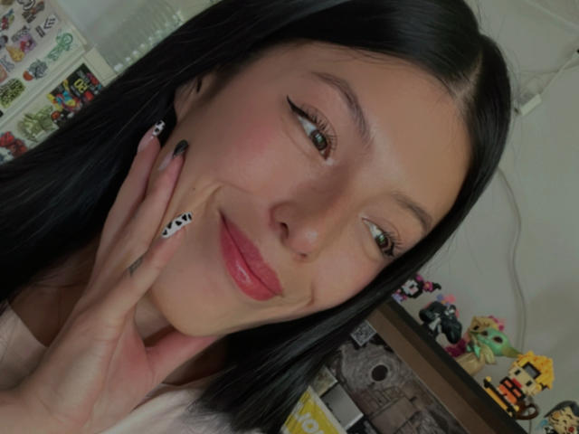 AizenMyoo has long black hair, a cute face with pink lips, and cow print nails, smiling at the camera with one hand on her cheek. She has a nose ring.