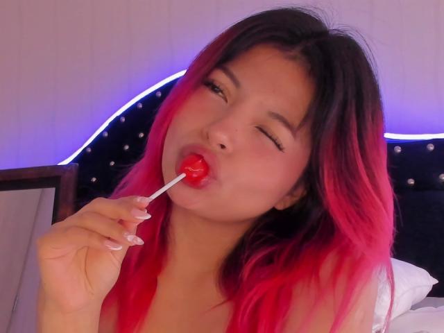 ShofiaWett with red and black hair, winking, sucking on a red lollipop, holding it with her right hand, showing her pink tongue. She's wearing a white top.