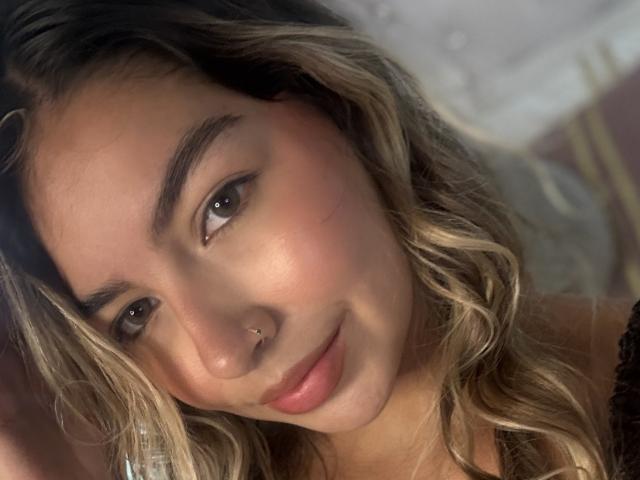 MaraCortez has wavy blonde hair, brown eyes, a nose ring, and a slight smile. Her skin is smooth, with a pinkish blush on her cheeks. She's wearing black lace.