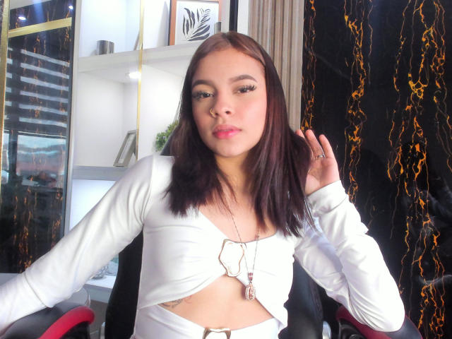 LunaFreya with brown hair, white crop top, showing her tits, hand in hair, necklace, sitting, chair, black and white wall, reflection.
