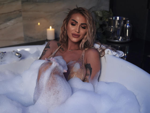 NicolSNaya with wet blonde hair, tattoos on arms, in a bubbly white bathtub, candle lit, looking down, relaxed, showing her tits, in a sensual, intimate setting.