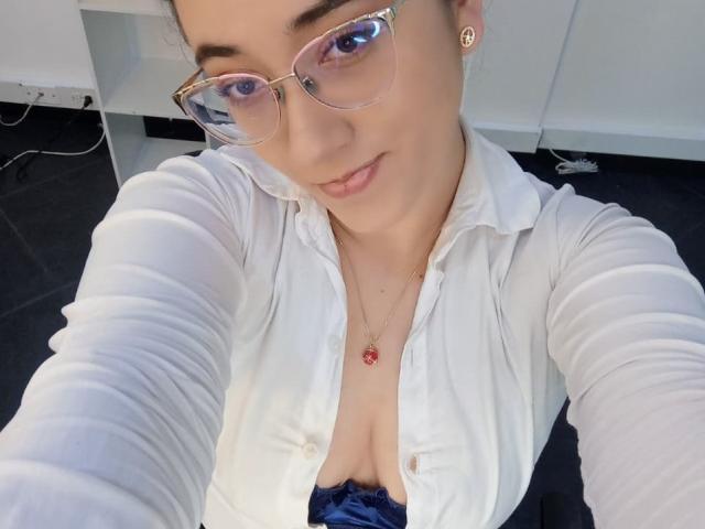 SwettieSammy with glasses, white button-up shirt, blue lace bra, showing her tits, smiling, gold necklace with red pendant, in office, holding phone for selfie.