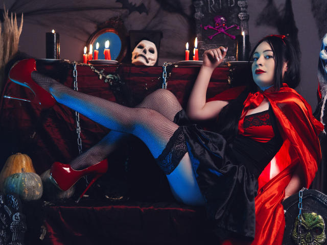 KristelRouse with black hair, red lipstick, black dress, red cape, fishnet stockings, and red heels, legs up, showing her ass, holding a cigarette, candles, skull, and bat.