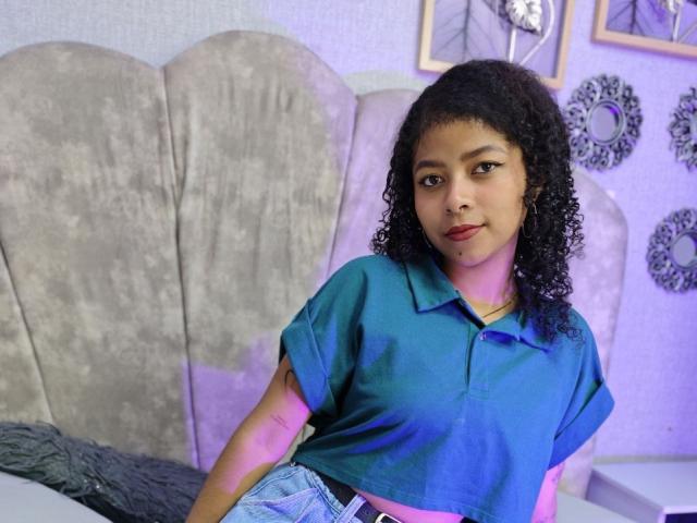 MistresSamanta with curly black hair, brown skin, and red lipstick, wearing a blue polo shirt and blue jeans, sitting on a gray upholstered chair, showing a slight smile.