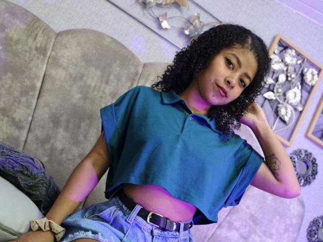 MistresSamanta with curly black hair, wearing a blue crop top and high-waisted jeans, has a tattoo on her right arm, sitting on a gray sofa, looking at the camera.