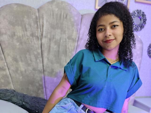 MistresSamanta with curly black hair, medium brown skin, wearing a blue polo shirt and denim jeans, smiling slightly, showing a small beauty mark on her chin.