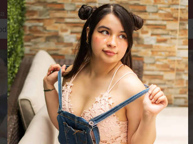 MiaNickole with double buns, pink lace bra, and denim overalls, holding straps, showing small tits, cute, freckled face, smiling, soft light, casual outdoor setting.