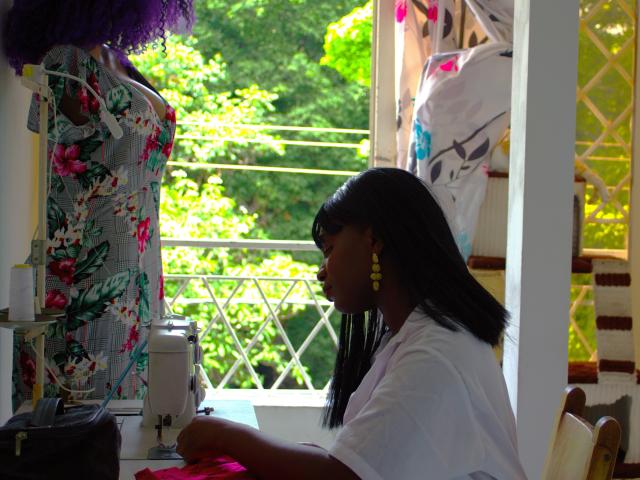 SaraBii with dark skin, straight black hair, yellow earrings, white shirt, sewing on purple fabric, floral dress mannequin, bright window, green trees outside, sunlit room, sewing machine.