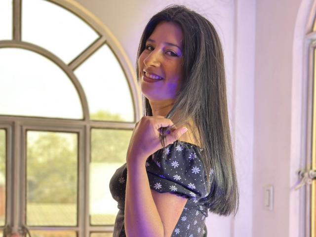 LuisaRojas with long black hair, smiling, holding a hair clip, wearing a black floral dress, purple eye makeup, standing by a window, looking over her shoulder.