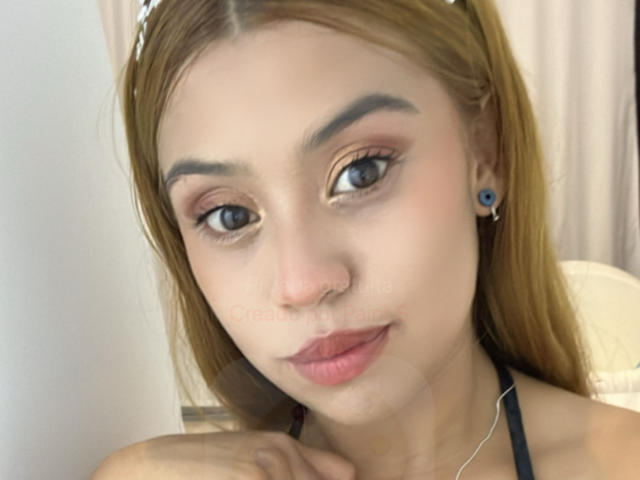 DakotaFenty tiene el cabello castaño claro, ojos azules y un piercing en la nariz. Lleva un top negro con tirantes, aretes azules y lápiz labial rosa, y está mirando a la cámara..