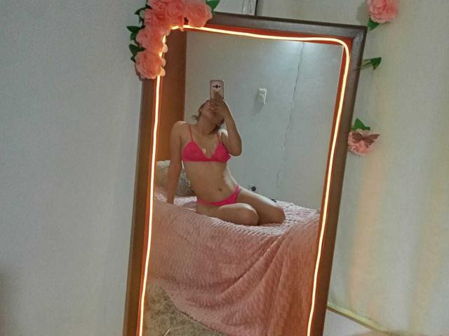 Mirror selfie of ZureyaAngels in pink lingerie, showing her tits and pussy, sitting on a pink bed, with glowing edge lights and pink flowers.