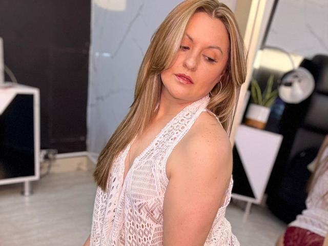 LaurenSpitz with blonde hair, wearing a white, crocheted sleeveless top, looking down with a slight smile, her small tits and pink lips visible, in a softly lit room.