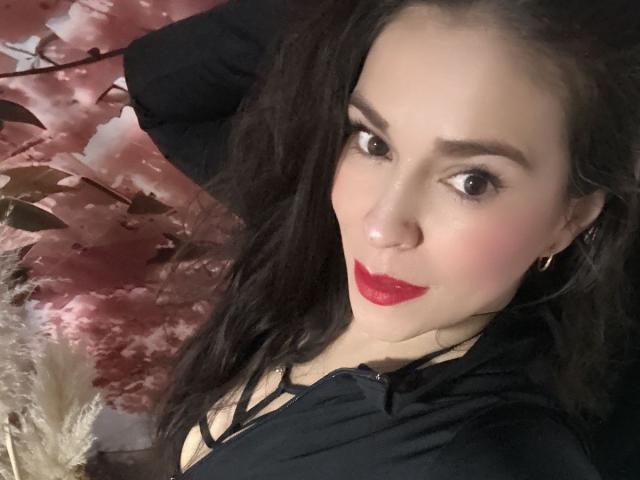 CelesteCorine mit dunklem Haar, rotem Lippenstift, schwarzem Oberteil und Reifenohrringen, die in die Kamera lächelt und in einer sexy, selbstbewussten Pose ihre Brüste zeigt..
