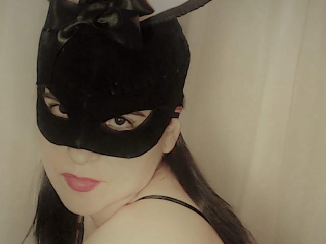 RossanaHairy with black cat mask, red lipstick, long black hair, and bare shoulders, looking seductively at the camera. Boobs partially visible, black straps on her shoulders.