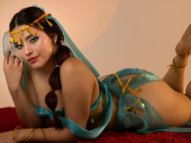 PamelaArabel lying on red carpet, wearing teal sheer lingerie with gold chains, her tits visible, long dark braid, gold headpiece, looking at camera with a slight smile.