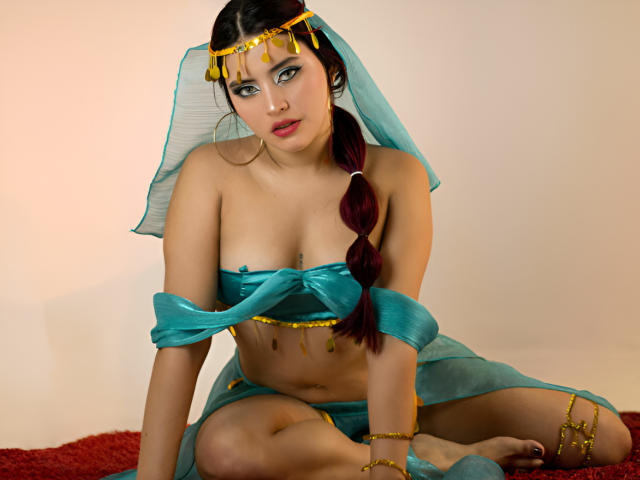 PamelaArabel with dark hair in a braid, wearing a teal headpiece and matching sheer outfit, exposing her tits and showing a gold chain on her arm.