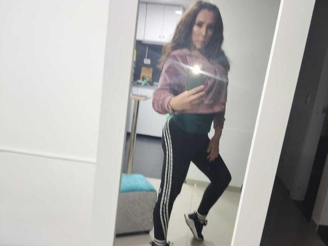 IssaMilfX with long brown hair, wearing a pink hoodie, green top, black Adidas track pants, and white socks, taking a mirror selfie, flashing a camera's flash.