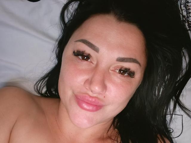 XCrazyBabeX has black hair, brown eyes, and is smiling with pink lips. She has thick eyebrows, long eyelashes, and a slightly flushed face, lying on a white bed.