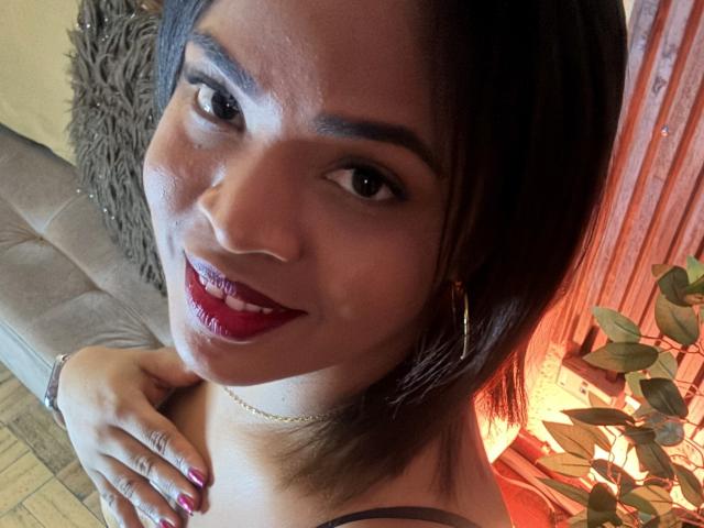 AliceBooty has a cute smile, red lipstick, and dark brown shoulder-length hair. She's wearing hoop earrings and a necklace, with her hand on her chest.