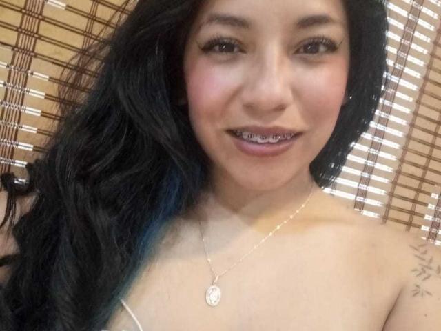 Paulina Colemann, con cabello negro largo, piel de color marrón claro y brackets metálicos, sonriendo, mostrando senos pequeños, llevando un collar de plata y un tatuaje en su brazo derecho..