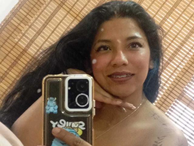 Paulina Colemann mit langem, schwarzem Haar, braunen Augen und kleinen, weißen Punkten im Gesicht, die ein Telefon mit einem blauen Aufkleber hält, lächelt und zeigt ihre nackten Schultern mit einem Tattoo..