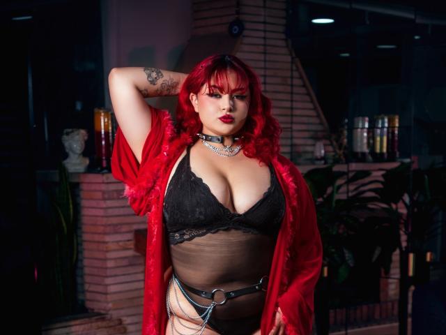 RavenVamp with red hair, black lace bra, and red fur robe, showing off her big tits and tattooed arm, wearing a chain collar and leather belt, in a dark room.