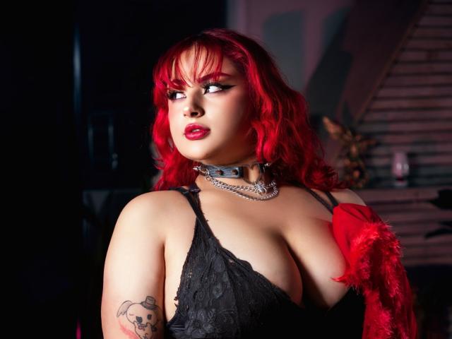 RavenVamp with red hair, black lace bra, red feather, silver chain, and tattoo, looking to the side with red lips. Boobs, pale skin, and sexy, goth vibe.