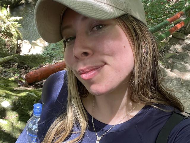 JennyLawry with light brown hair, green eyes, and a subtle smile, wearing a beige cap, navy shirt, and a necklace, has a bottle on her right.