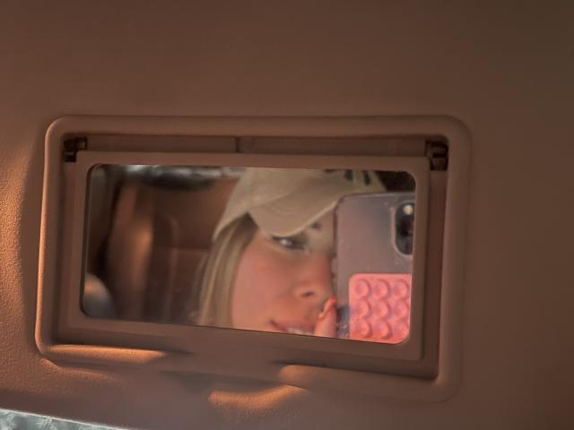 JennyLawry's face in a car mirror, wearing a beige hat, with blonde hair, holding a pink phone case, reflecting in a rectangular mirror with a beige frame.