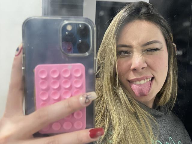JennyLawry with blonde hair, sticking out tongue, winking, holding phone with pink case, showing nails, in selfie. Phone has three cameras, her tits not visible.
