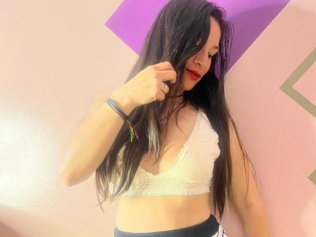 AlannaFox, com cabelos longos e pretos, um top de renda branco, batom vermelho e shorts pretos, tocando o cabelo, usando uma pulseira colorida, em um quarto rosa..