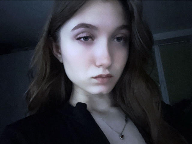 SilverxKitty has pale skin, long brown hair, and a serious expression. She's wearing a black top with a silver necklace and has subtle makeup with pink eyeliner.