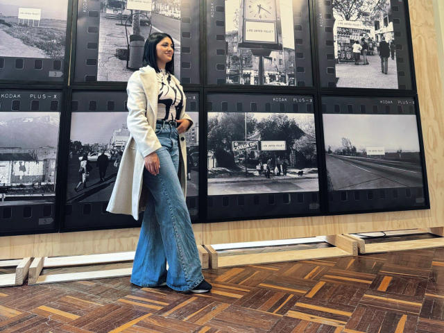 MaddieDemie with black hair, white coat, black-and-white striped shirt, blue flared jeans, standing on wooden floor, in front of black-and-white film strip wall, looking to side.