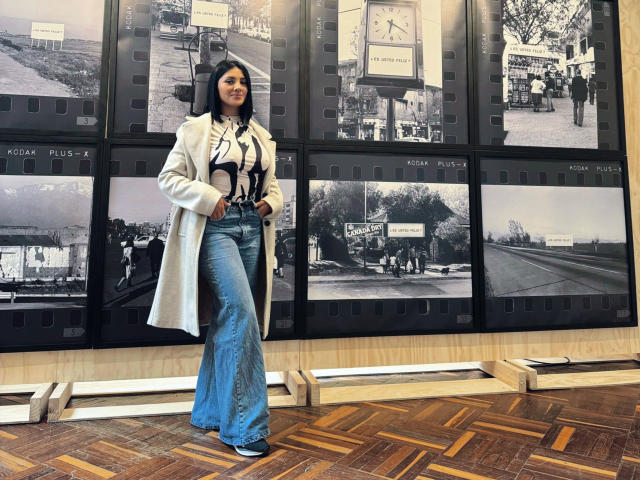 MaddieDemie with long black hair, wearing a white coat, black strappy top, and blue flared jeans, standing in front of black-and-white photo displays, hands in pockets.