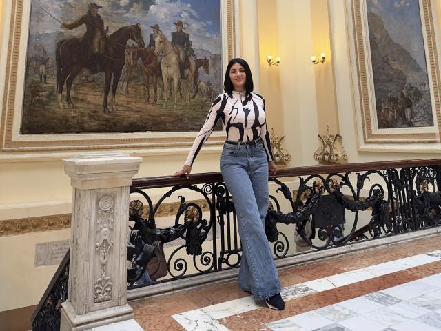 MaddieDemie stands with ass slightly raised, wearing a striped top, high-waisted blue jeans, and black shoes, leaning on an ornate black metal railing in a fancy room.