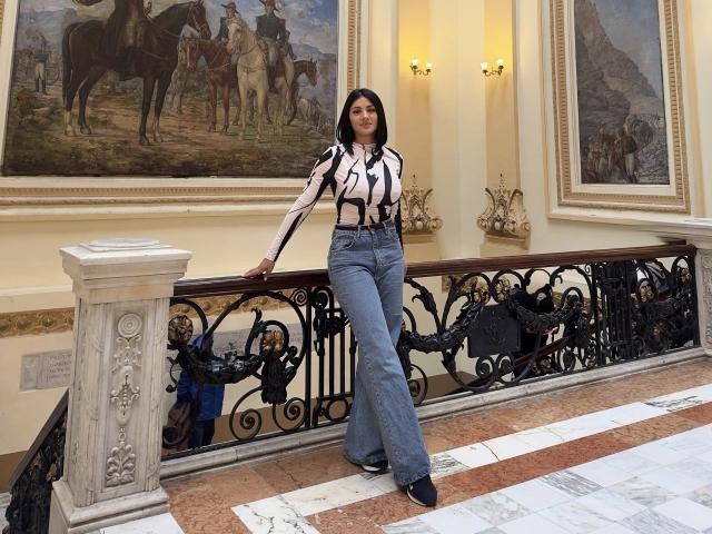 MaddieDemie with tits, wearing a striped shirt, black suspenders, and blue flared jeans, standing against a black iron railing, marble floor, and wall paintings.