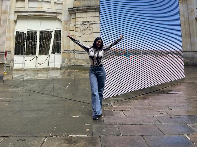 MaddieDemie stands with arms out, wearing a sheer black top, high-waisted blue jeans, and black heels, in front of a blue shuttered building, looking confident.